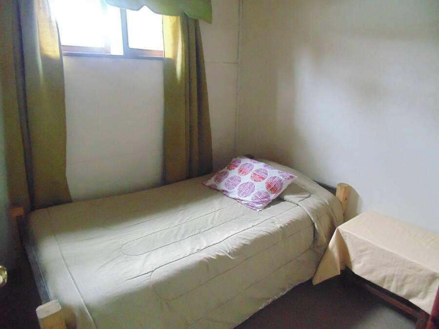Een kleine kamer met een enkel bed, een kussen, een tafel met tafelkleed en een raam met gordijnen.