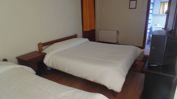 A room with two single beds, a wooden nightstand, a desk with a chair, a radiator, and a television.