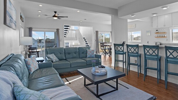 A spacious living room with a grey sofa, a coffee table, and a kitchen area with bar stools and white cabinets.