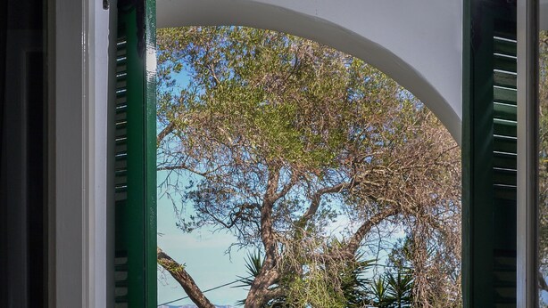 Puerta abierta a un balcón con mesa y sillas, un vaso de zumo de naranja y vistas a árboles y al mar.