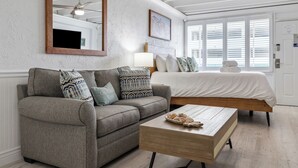 A modern bedroom with a gray sofa, a wooden coffee table, a bed with white bedding, a ceiling fan, and a large window with blinds.