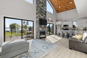 A spacious living area with a fireplace, a large window overlooking a deck, and a kitchen with modern appliances. - Newer Cottage on all sports lake, pontoon boat available (Trufant)