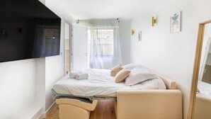 A single bed with white bedding, a bedside table, a chair, a window with sheer curtains, and a wall-mounted TV. - Lavie Maison: Equipped Studio, Chartrons (Bordeaux)