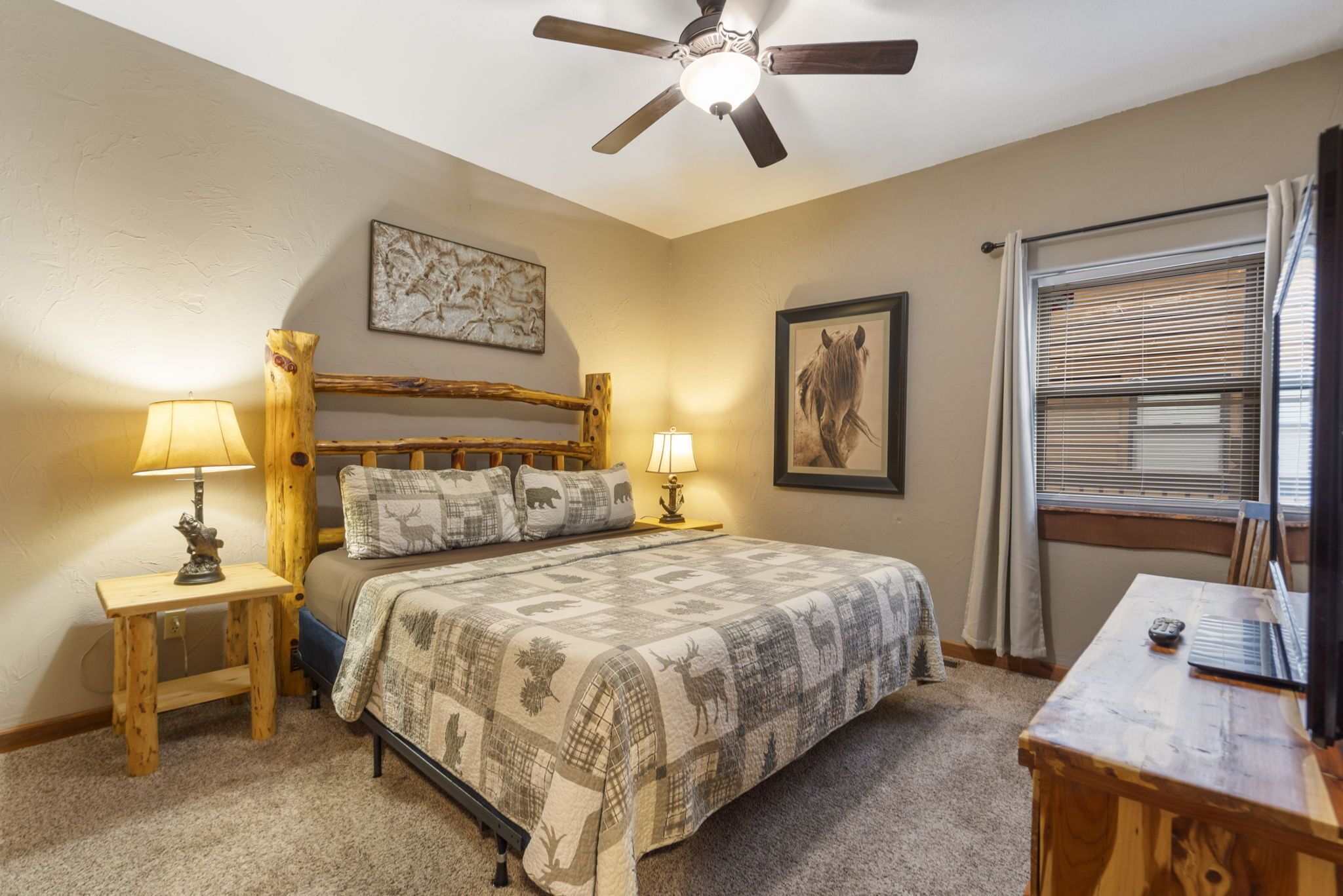 A bedroom with a bed, two bedside tables with lamps, a ceiling fan, a window with blinds, and a framed picture on the wall.