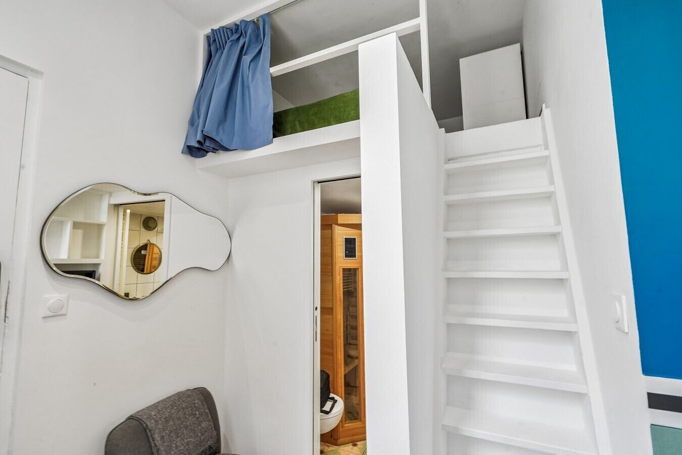 A room with a white wall, a mirror, a chair, a blue curtain, and a staircase leading to a loft area.