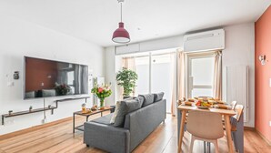 A modern living room with a flat-screen TV, grey sofa, dining table with chairs, and a red pendant light.