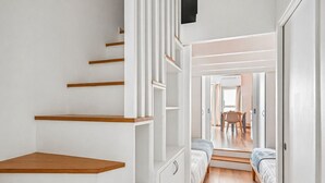 A narrow hallway with a staircase, a bed, and a dining area visible through an open door.