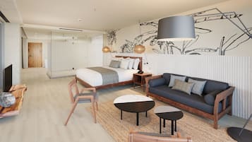 Una habitación de hotel moderna con una cama grande, un sofá, una mesita y una silla. Se aprecia un mural en la pared y un ventanal amplio.