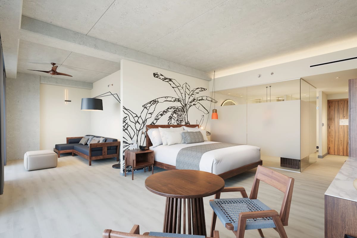 A modern hotel room with a large bed, a sofa, a dining table with chairs, and a ceiling fan.
