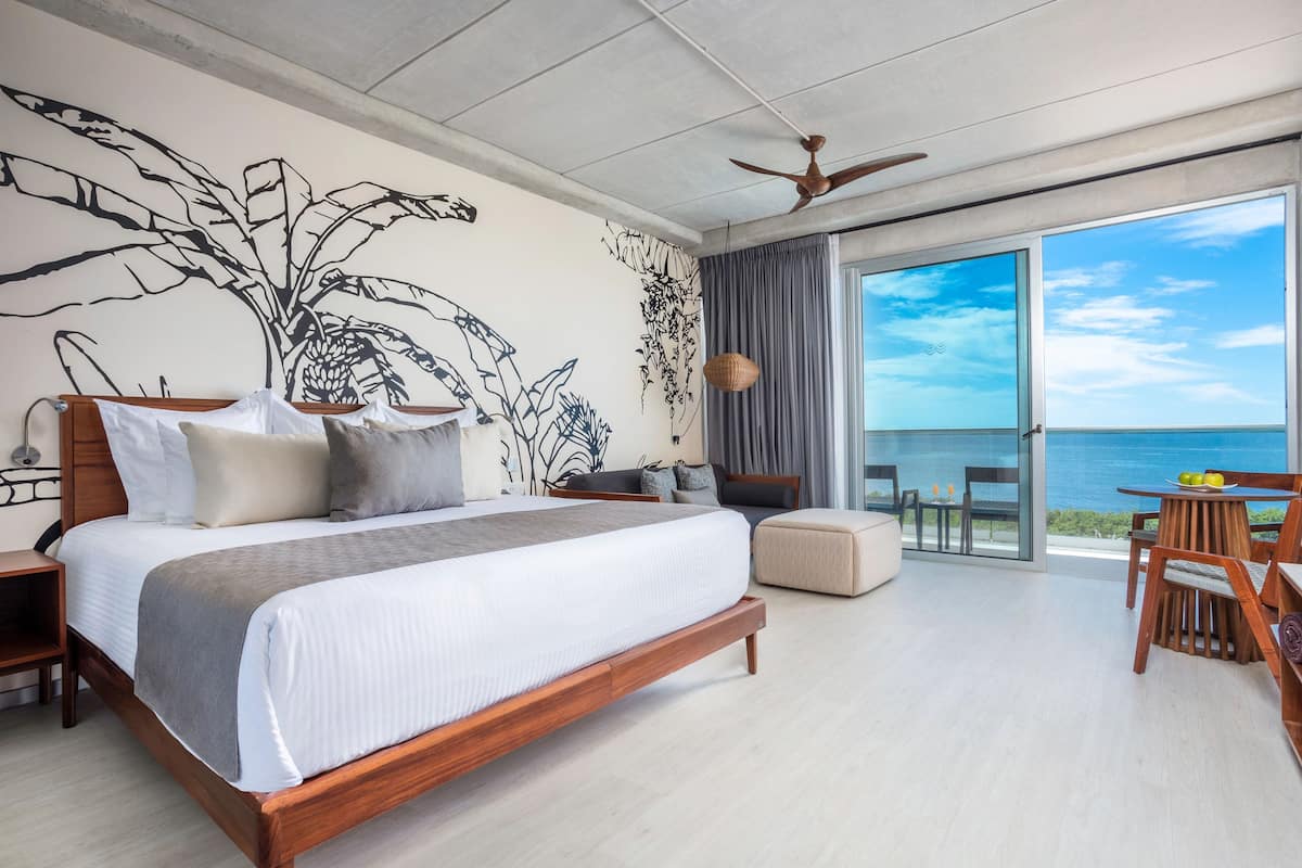 A modern bedroom with a large bed, a ceiling fan, a view of the ocean, and a balcony with a table and chairs.