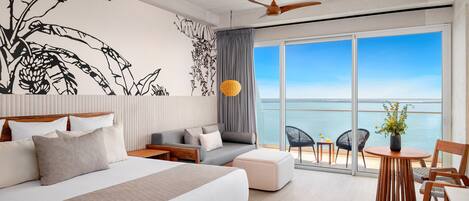 Una habitación de hotel moderna con una cama grande, un sofá, un balcón con vistas al mar y un ventilador de techo.