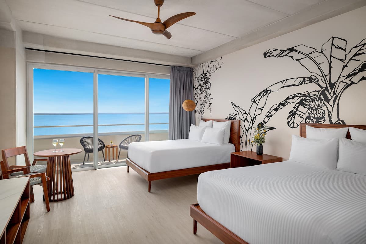 A hotel room with two beds, a balcony with a view of the sea, a ceiling fan, and a wall mural.