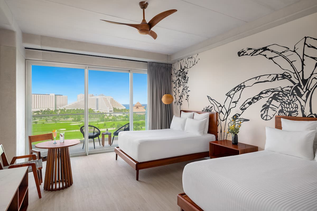 A hotel room with a large bed, a smaller bed, a dining table, and a chair. There is a large window with a view of buildings and a green area.