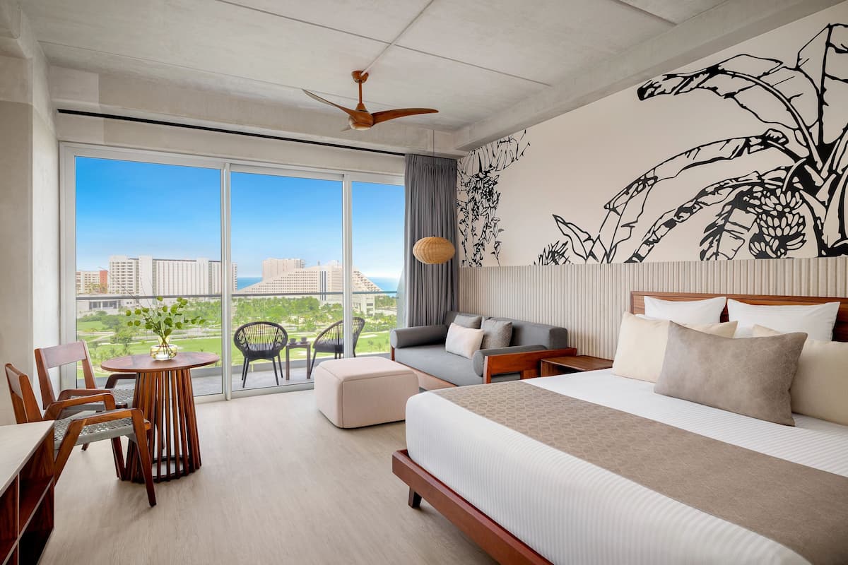 A hotel room with a large bed, a desk with a chair, a small table, and a balcony with a view of buildings and greenery.