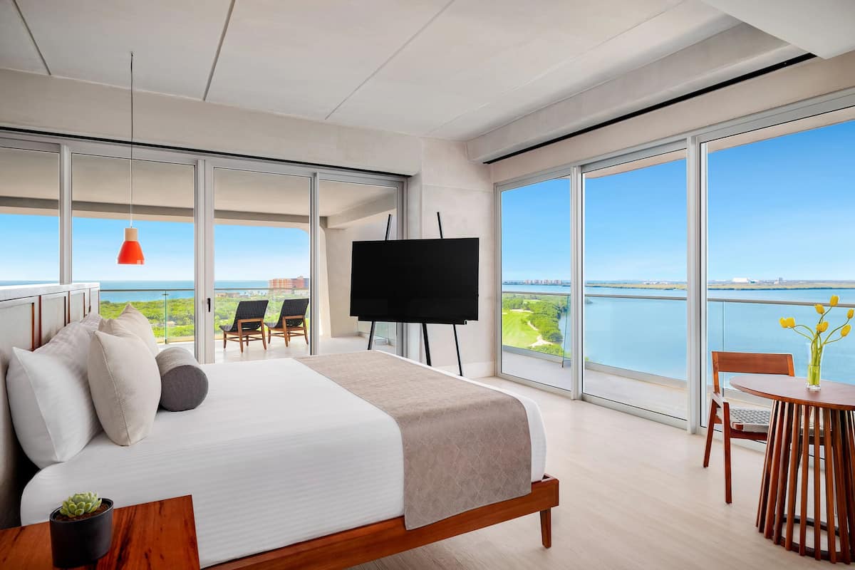 A modern hotel room with a large bed, a flat-screen TV, a dining area, and a balcony with a view of the water.