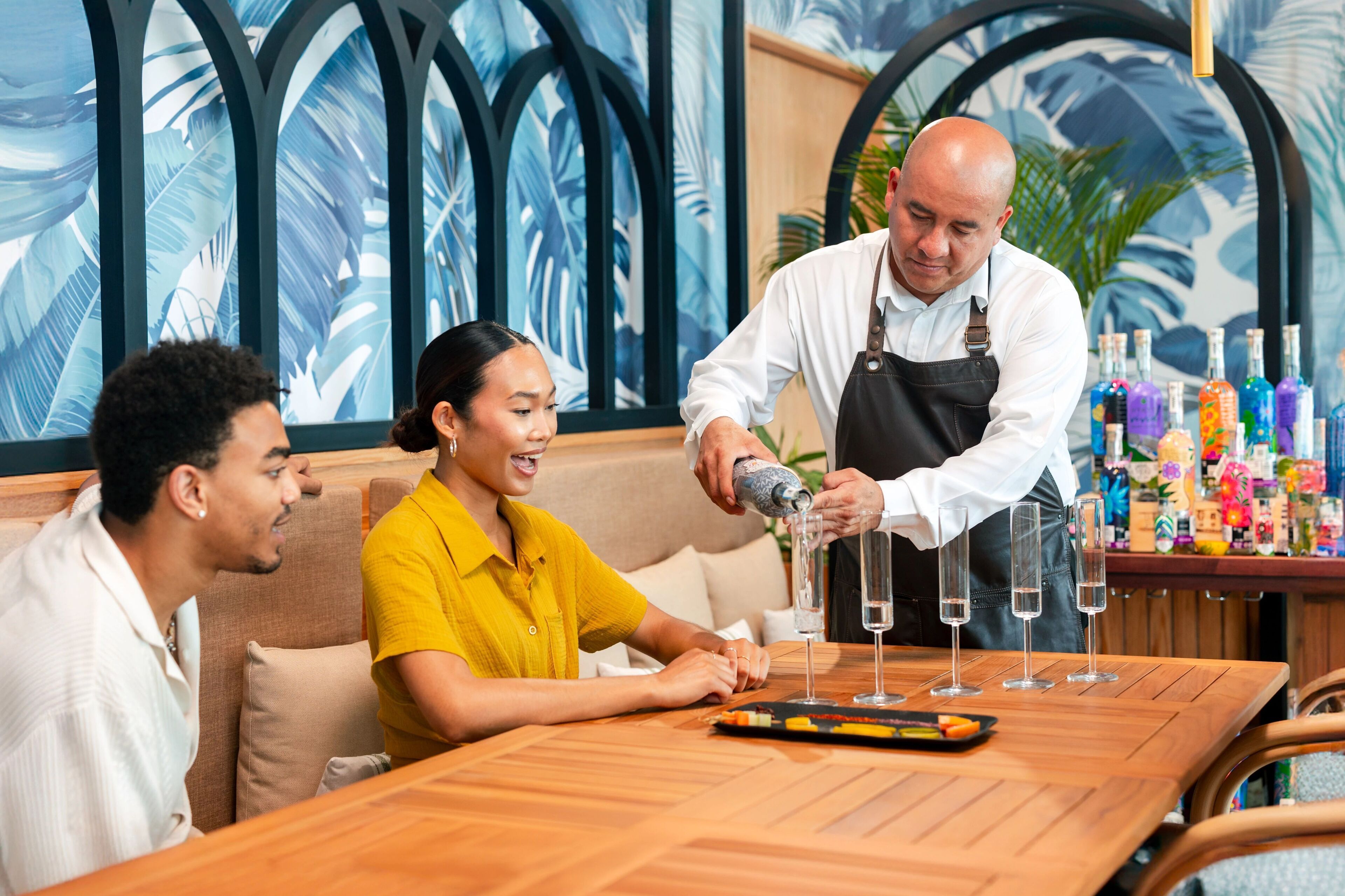 A bar setting with a bartender preparing drinks. There are three people seated at a wooden table with a tray of snacks. The background features a tropical mural with palm leaves.