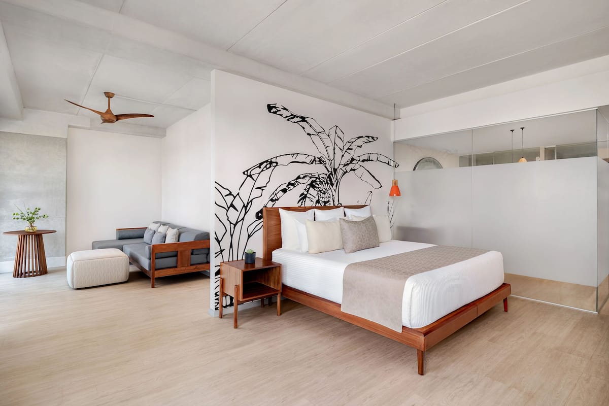 A modern bedroom with a large bed, a wooden nightstand, a grey sofa, and a ceiling fan.