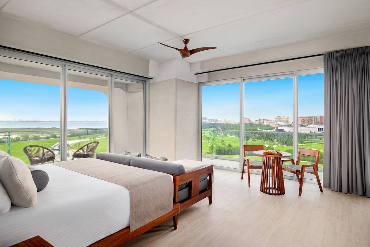 A spacious bedroom with a large bed, a dining table, and chairs, a ceiling fan, and a view of a golf course and city skyline through large windows.