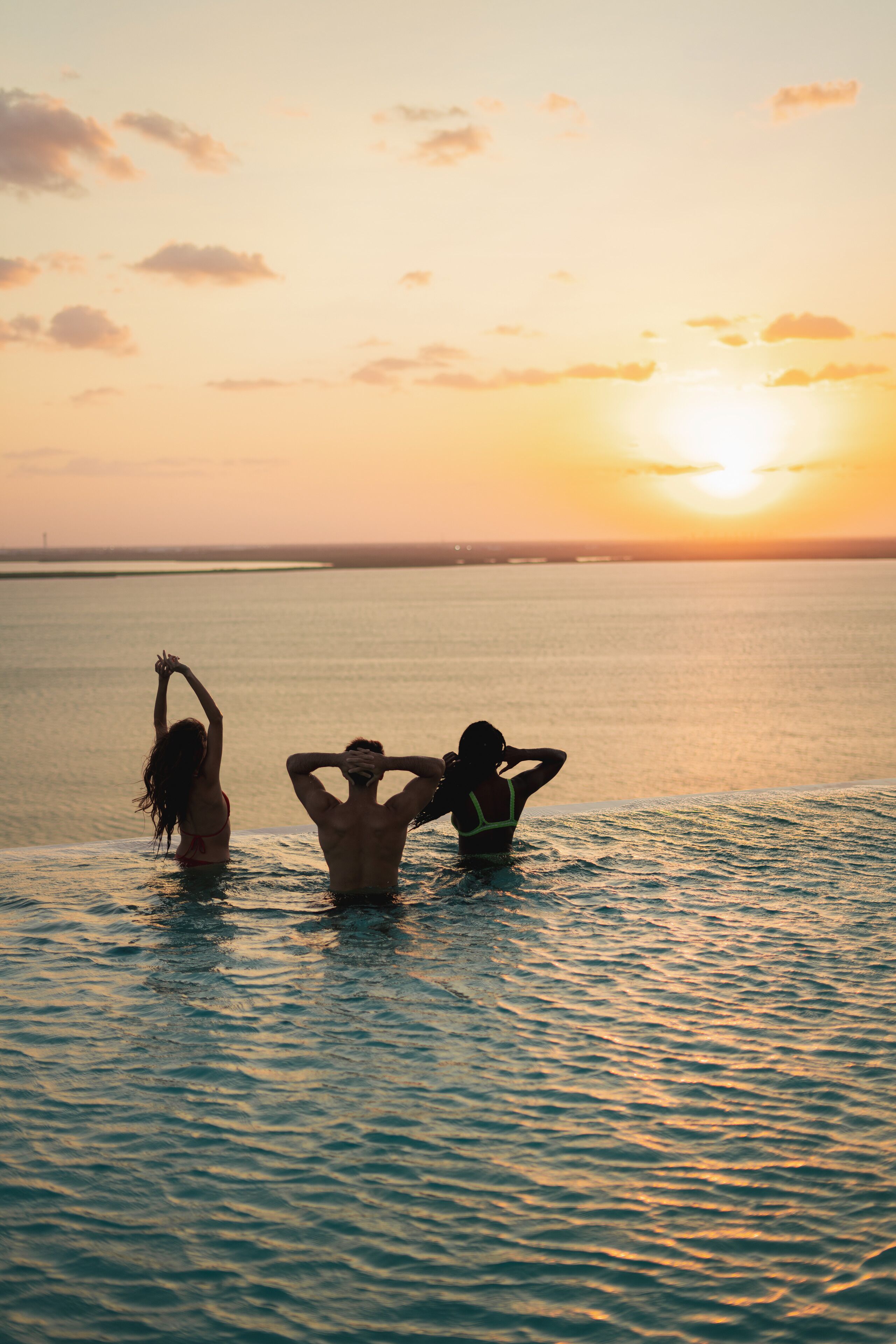 Un atardecer sobre un mar tranquilo con tres figuras siluetas en el agua.