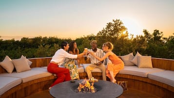 A group of people gathered around a fire pit on a wooden deck with cushioned seating.