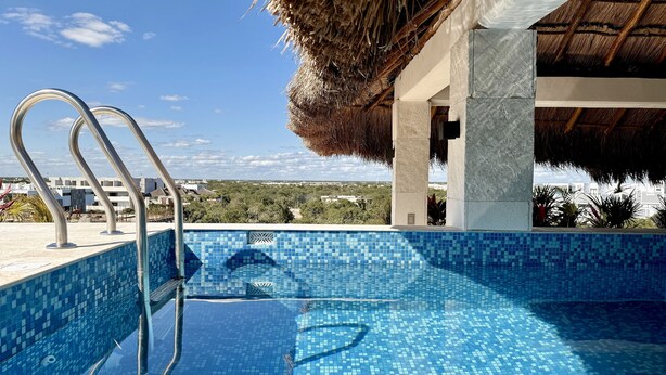 A swimming pool with a metal ladder, surrounded by a tiled deck and a thatched roof structure.