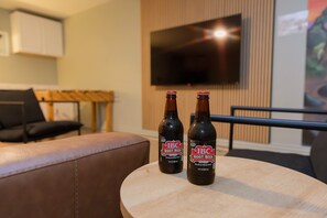 To flasker IBC Root Beer på et træbord i et rum med en lædersofa, et fjernsyn monteret på væggen og en træstol.