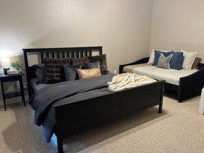 Un dormitorio con una cama negra, una mesita de noche con lámpara, una planta pequeña y una silla con una manta a cuadros.