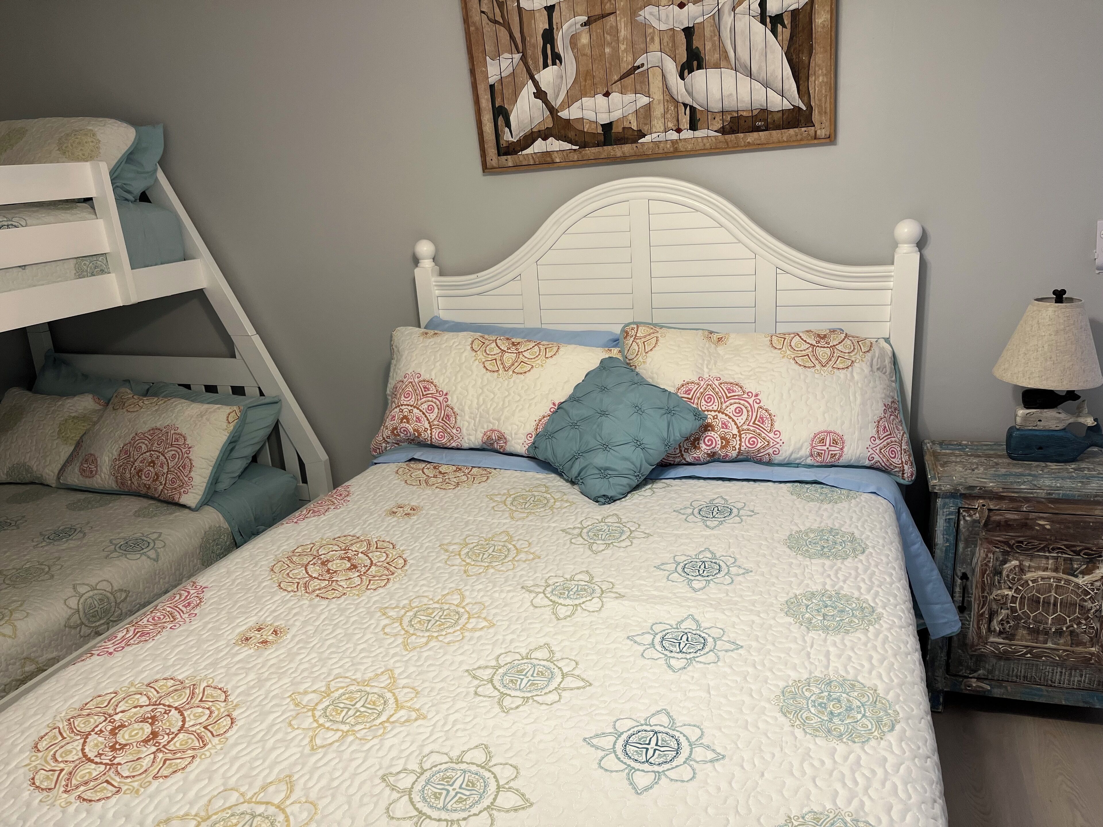 Um quarto com uma cama de solteiro, uma cama com roupa de cama estampada, uma pintura de pássaros na parede e uma luminária em uma mesa de cabeceira.