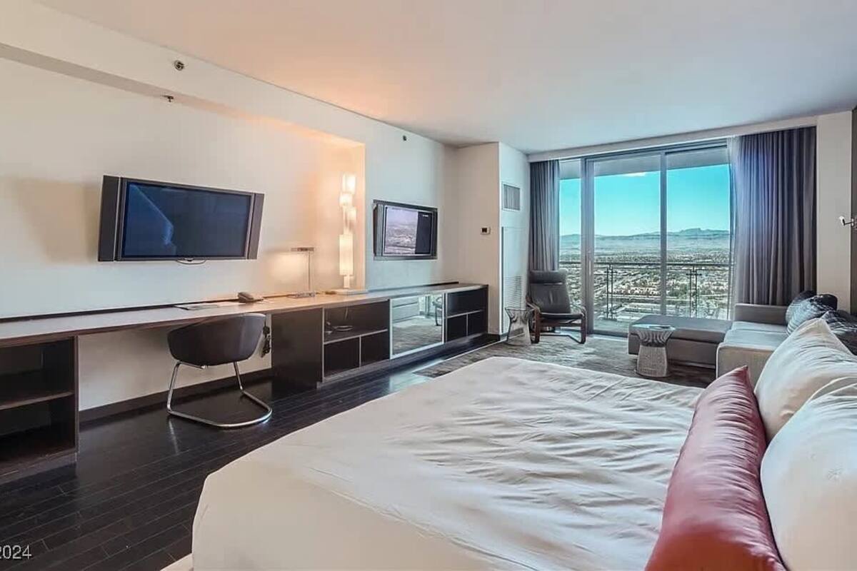 A modern hotel room with a large bed, a desk with a chair, a TV, and a balcony with a city view.