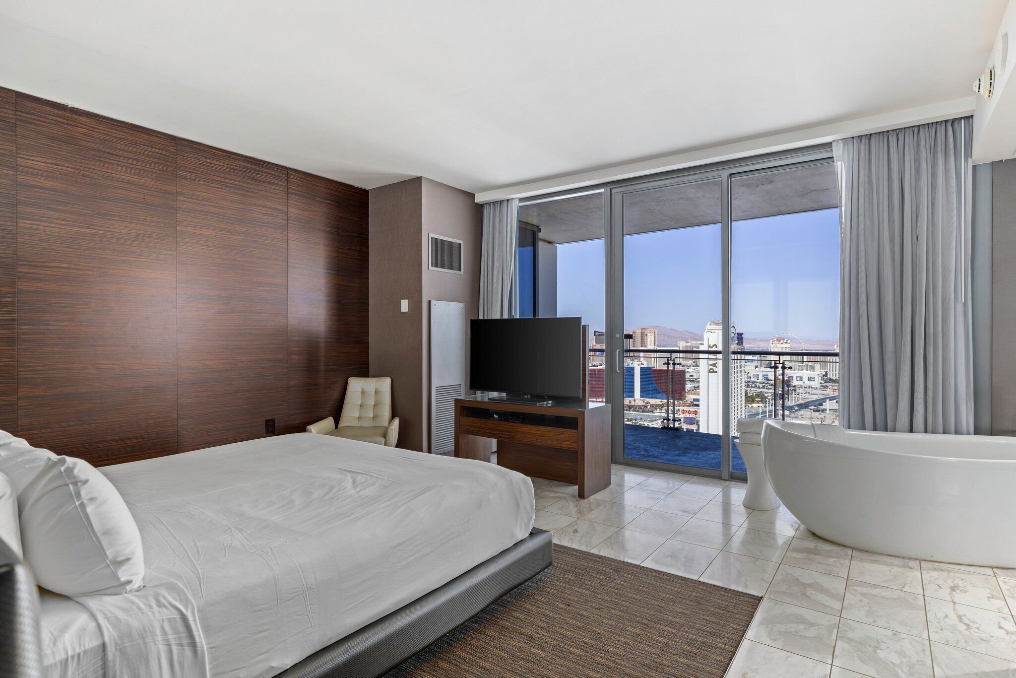 Een moderne hotelkamer met een groot bed, een flatscreen televisie, een bank, een badkuip en een balkon met uitzicht op de stad.