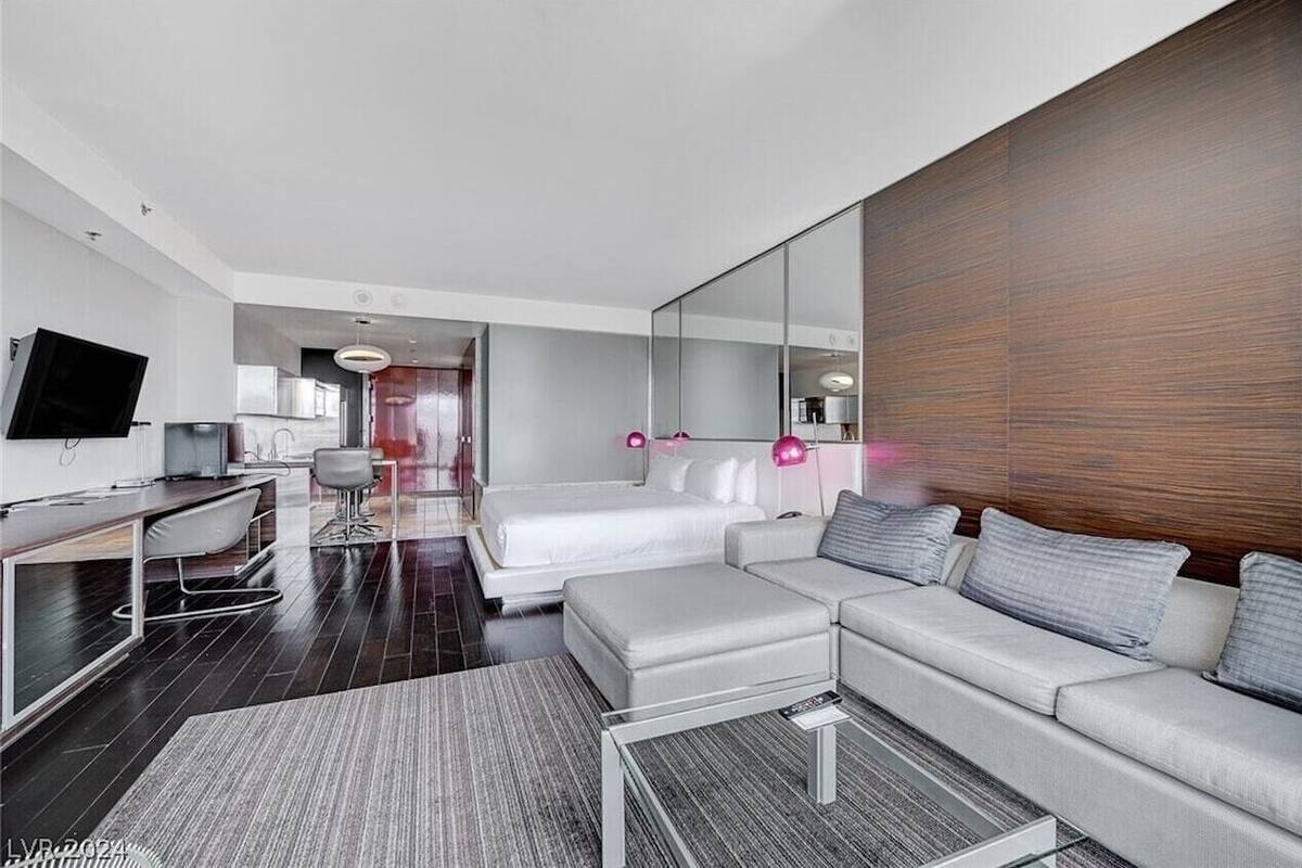 A modern hotel room with a large bed, a sofa, a desk with a chair, a TV, and a large mirror.