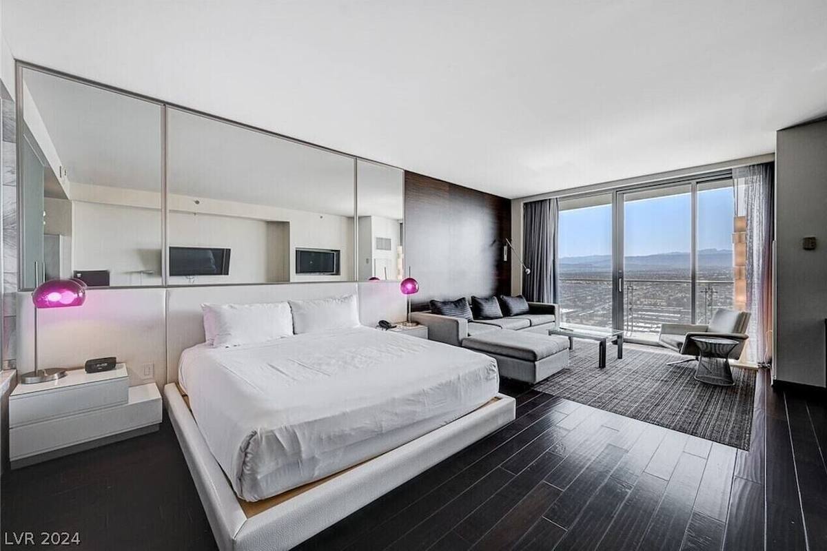A modern hotel room with a large bed, a sofa, a chair, and a large window offering a city view.