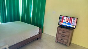 Een slaapkamer met een bed, een houten nachtkastje, een televisie waarop een nieuwsprogramma draait, en groene gordijnen.