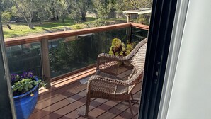 A balcony with a wicker chair, a blue planter with flowers, and a view of a garden with trees and a fence.