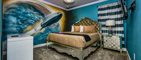 A bedroom with a bed, a dresser, a nightstand, a lamp, and a wall mural.