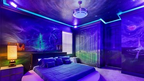 Un dormitorio con una cama centrada sobre una plataforma circular, una mesita de noche con lámpara, un armario y una pared con un mural con temática espacial.