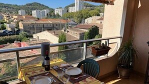 A balcony with a table set for two, a bottle of wine, and a glass. The balcony overlooks a cityscape with buildings and greenery.