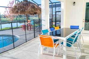 A modern outdoor dining area with a glass table and chairs, a thatched umbrella, and a view of a lake and greenery. - Sports Bowling Home, Orlando Mansion 6229 (Orlando)