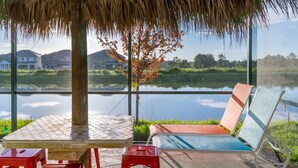 A patio with a table and chairs, a thatched roof, and a view of a lake and buildings in the distance.