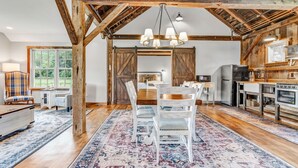 Un interior rústico con vigas de madera a la vista, una zona de comedor con sillas blancas, una cocina con estufa y un ventanal grande.