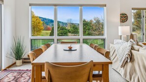 A dining room with a large table, chairs, and a view of a forested landscape through large windows.