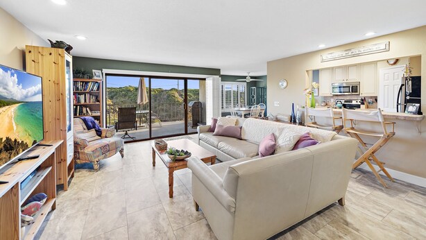 Een ruime woonkamer met een grote bank, een televisie en een balkon met uitzicht op een strand.