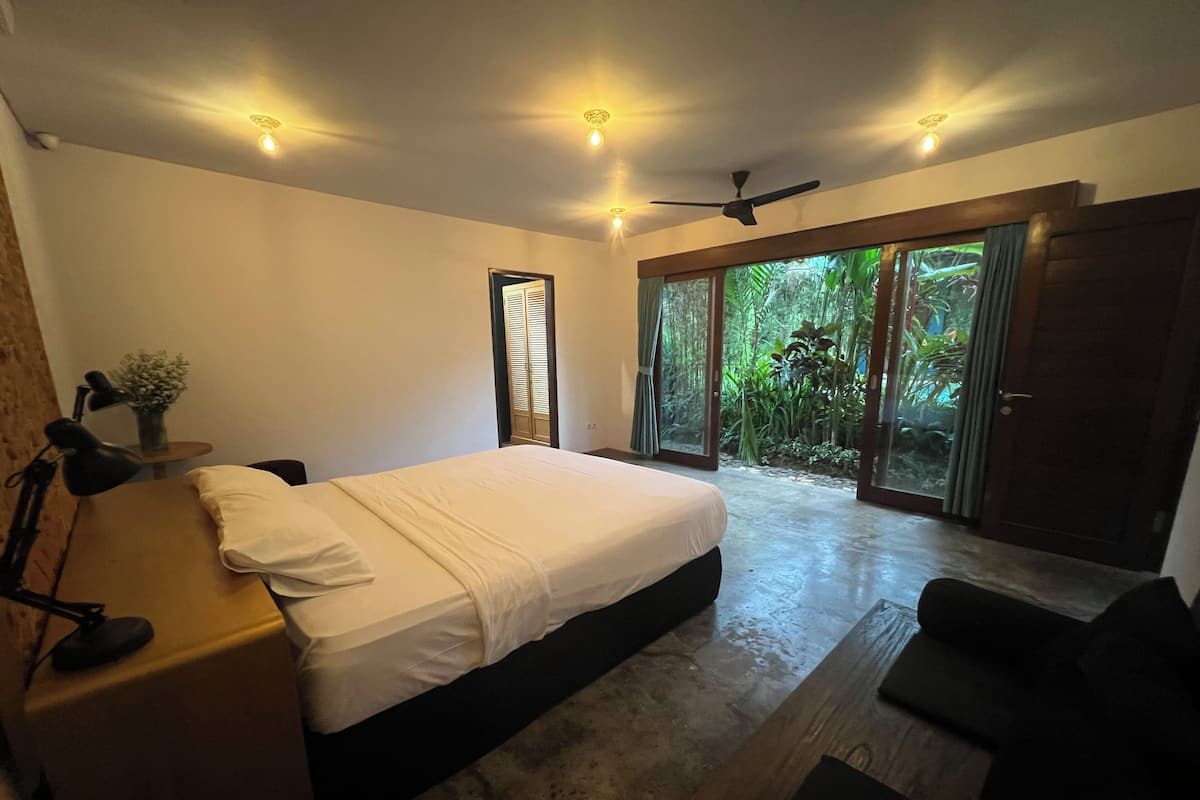 A bedroom with a bed, a desk, a chair, a ceiling fan, and a view of greenery through a sliding door.