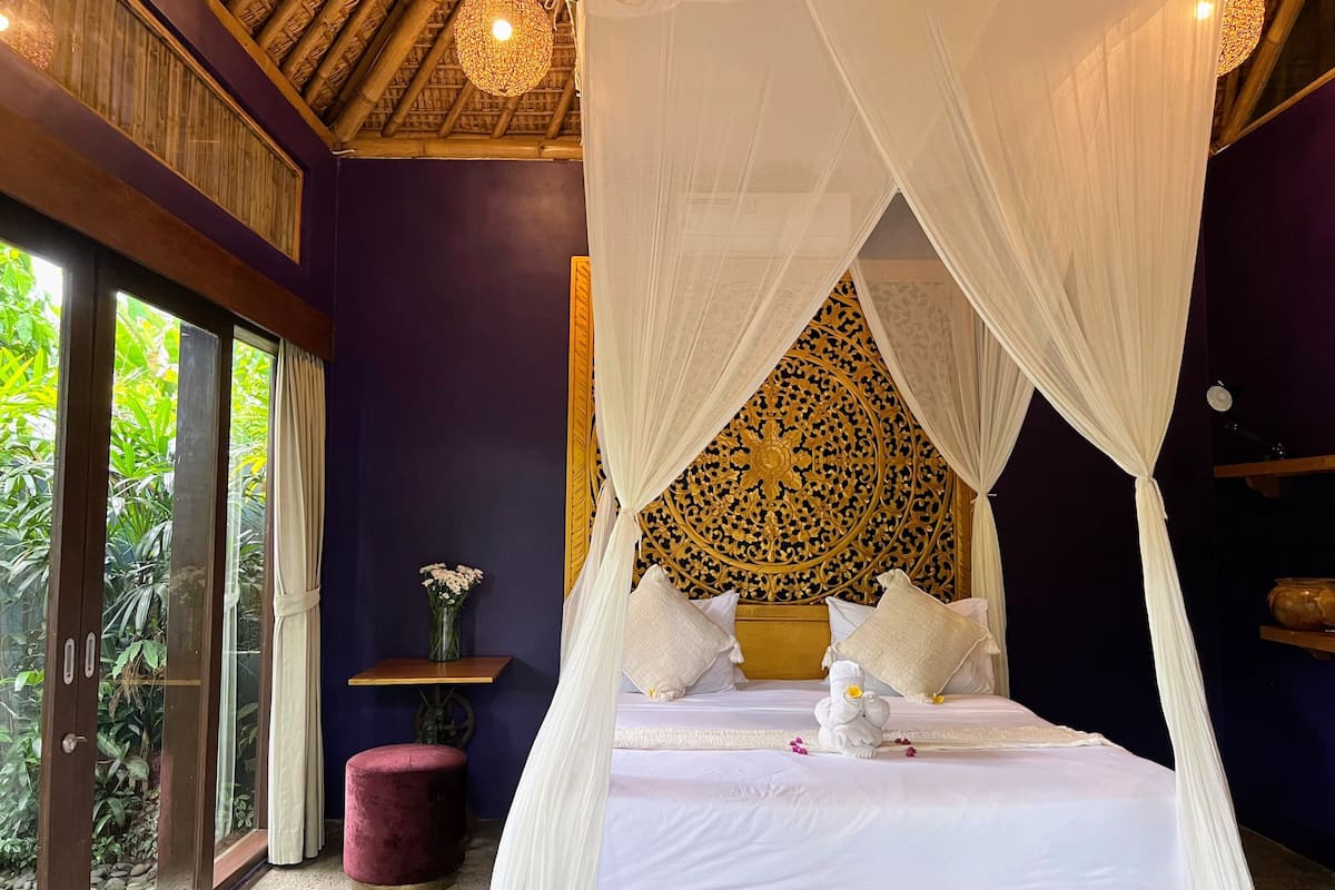 A room with a bed under a canopy, a small table with a vase, and a view of greenery outside.