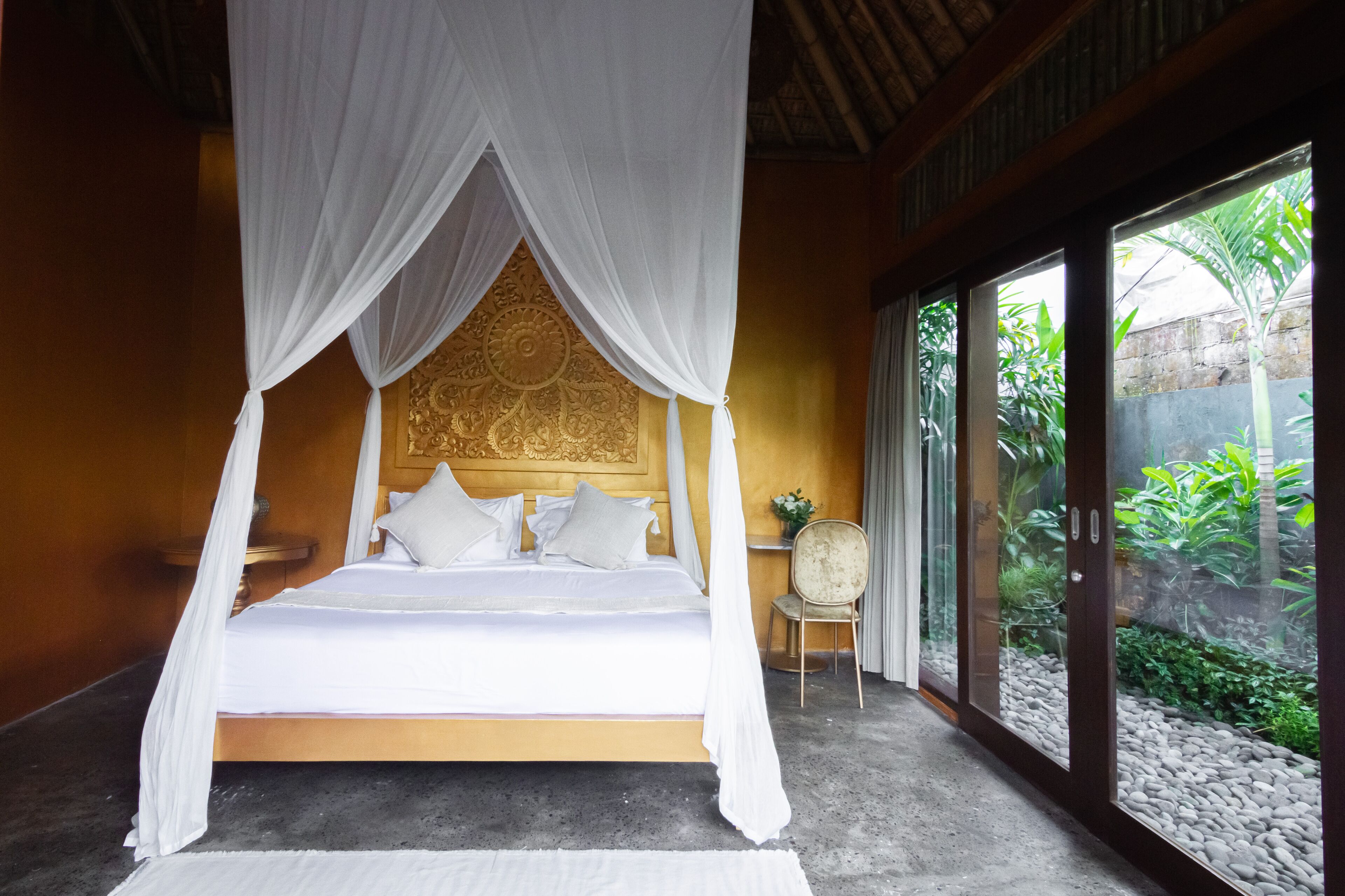 A bed with a canopy, a chair, and a view of greenery through large windows.