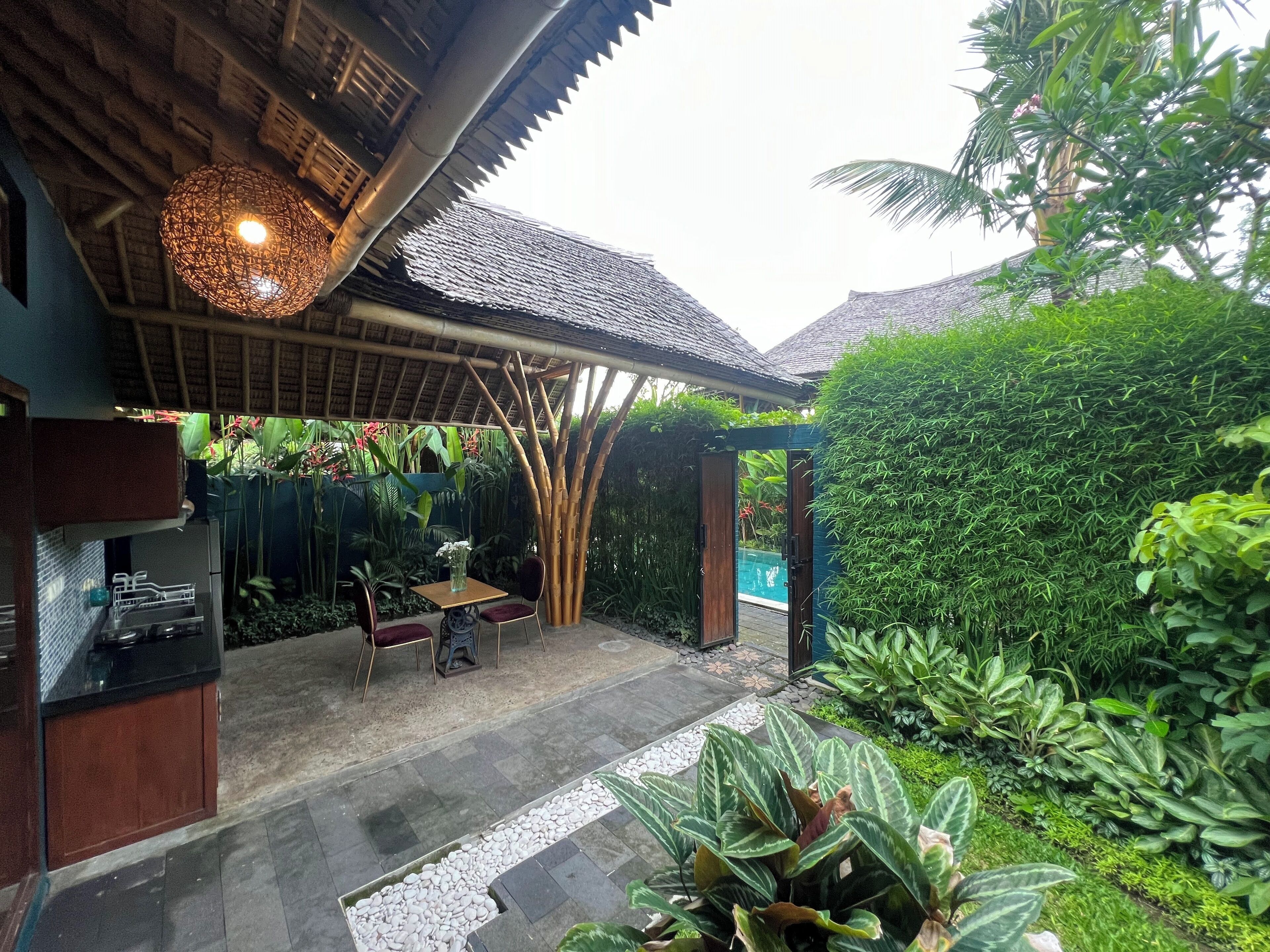 A covered outdoor seating area with a table and chairs, surrounded by lush greenery and a bamboo structure.