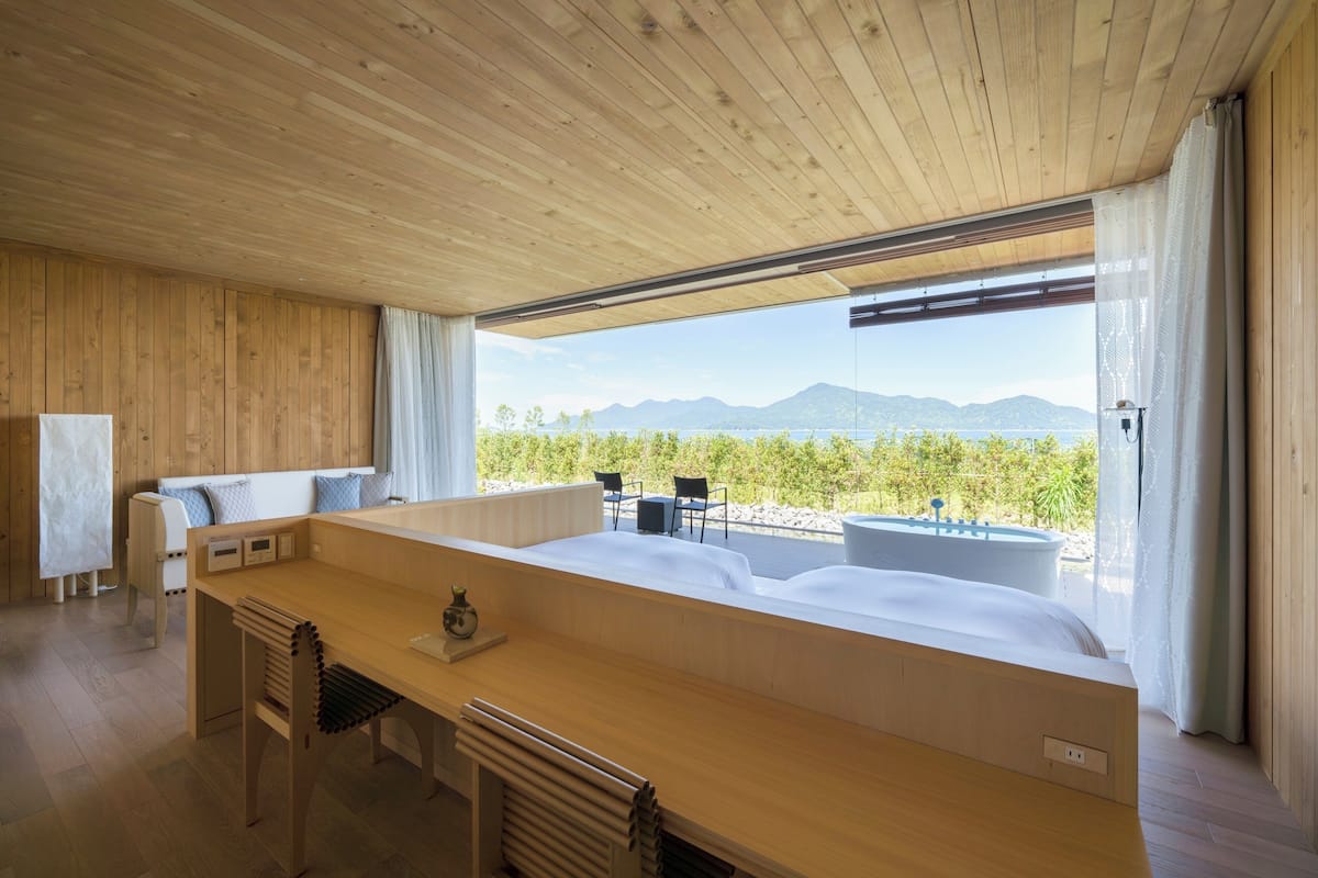 A modern room with a wooden ceiling, a large bed, a wooden desk, and a view of mountains and greenery.