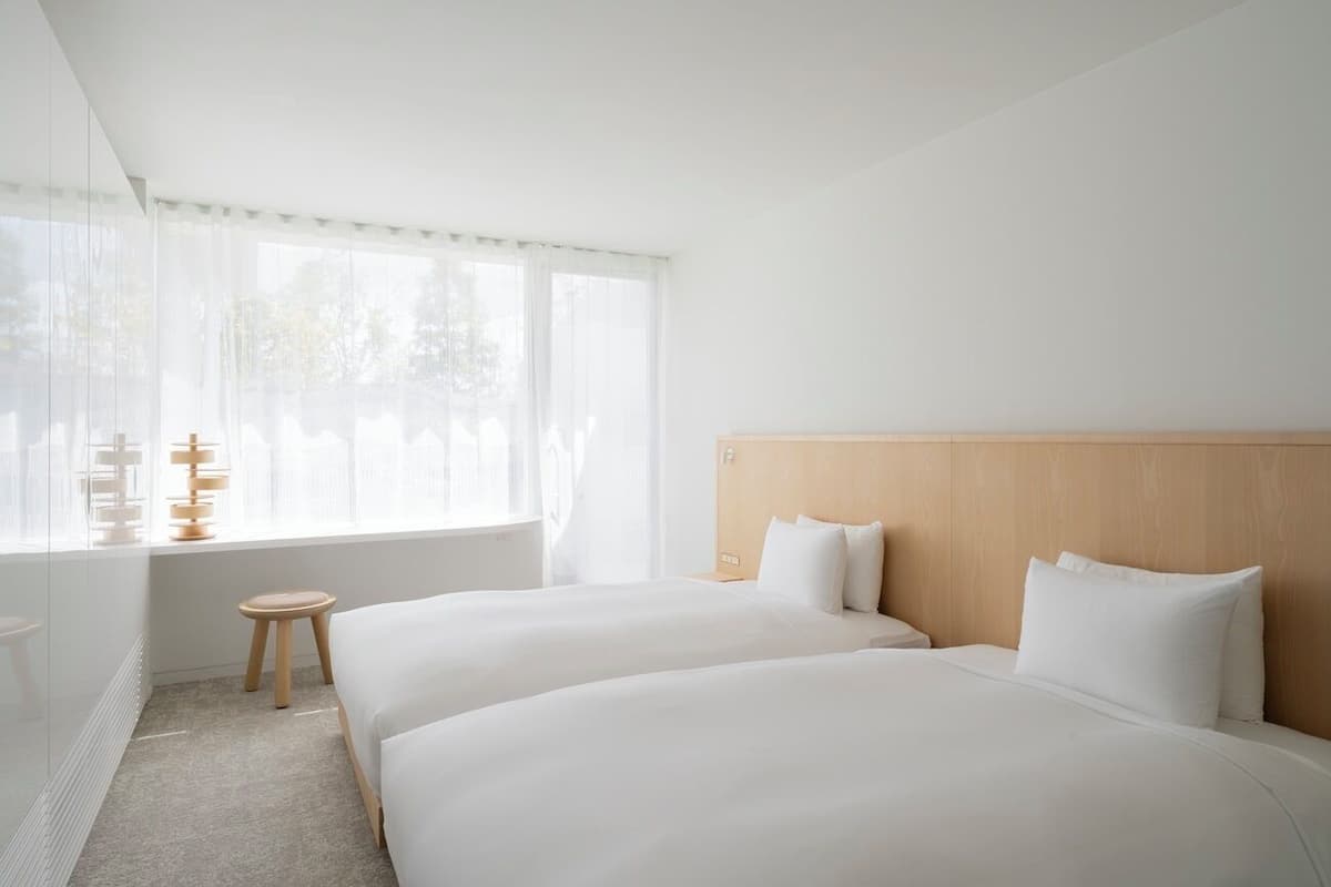 A hotel room with two beds, a wooden headboard, a small stool, and a window with sheer curtains.