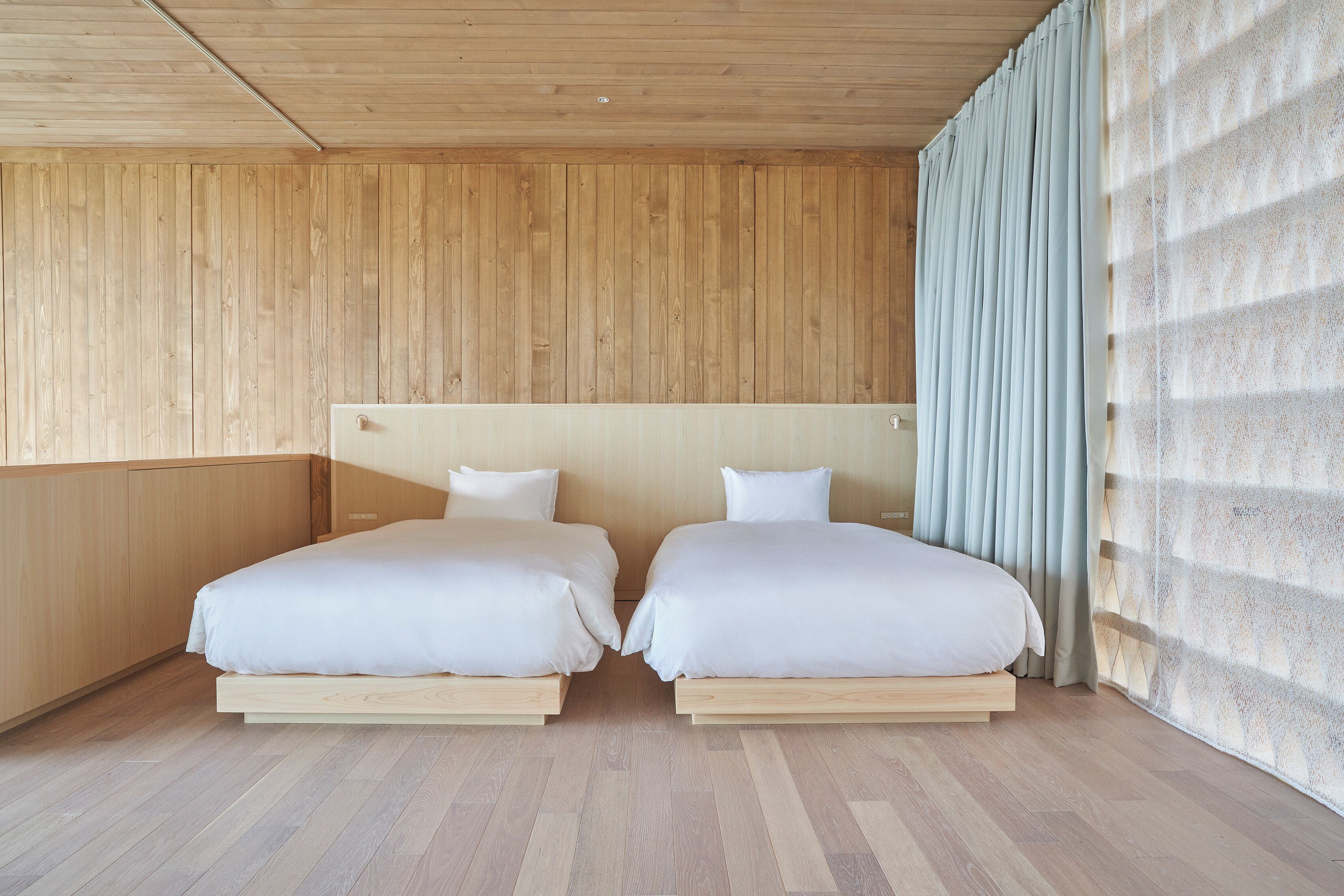 Una habitación con dos camas, paredes de madera y un ventanal con cortinas.