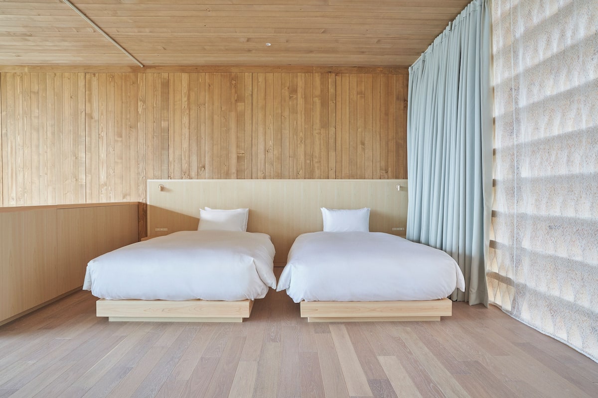 A room with two beds, wooden walls, and a large window with curtains.